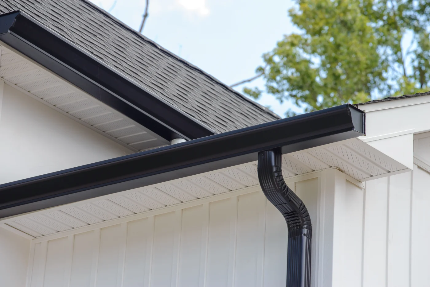 A close-up view of black gutters and a black downspout on a white house with an asphalt shingle roof.