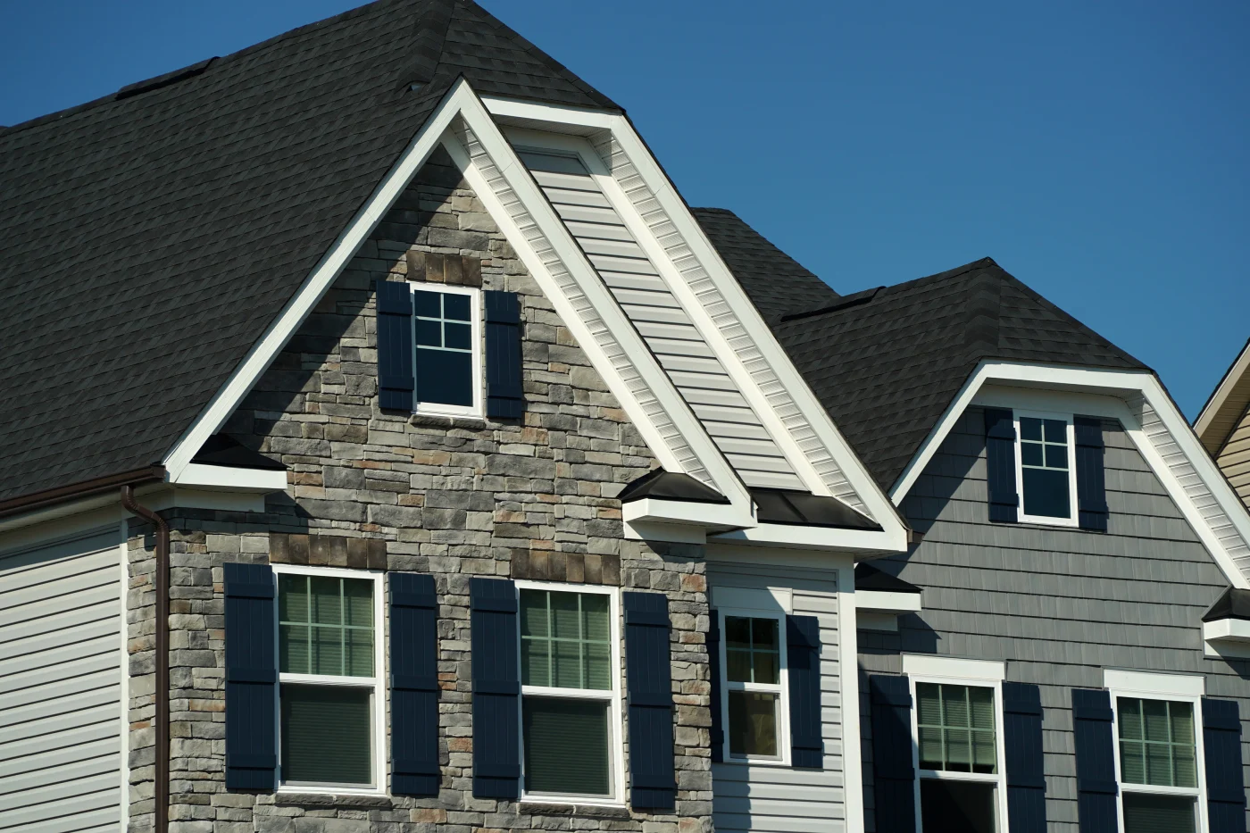 A white, gray, and stone house with white windows, blue shutters, and a dark asphalt shingle roof.