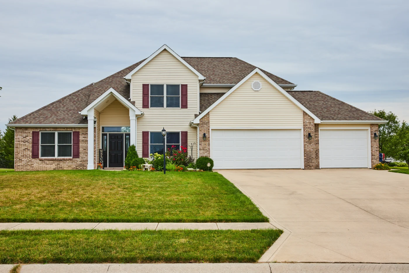 A yellow and brick house with two white garage doors, white windows with brown shutters, a brown front door, and an asphalt shingle roof.