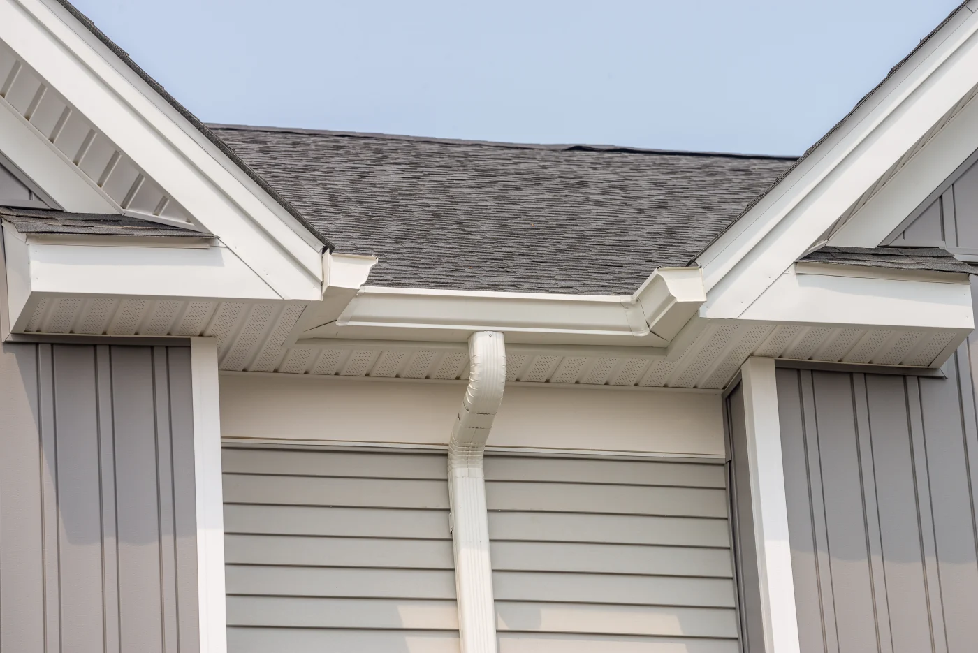 White gutters and a white downspout on a gray house with white trim and an asphalt shingle roof.