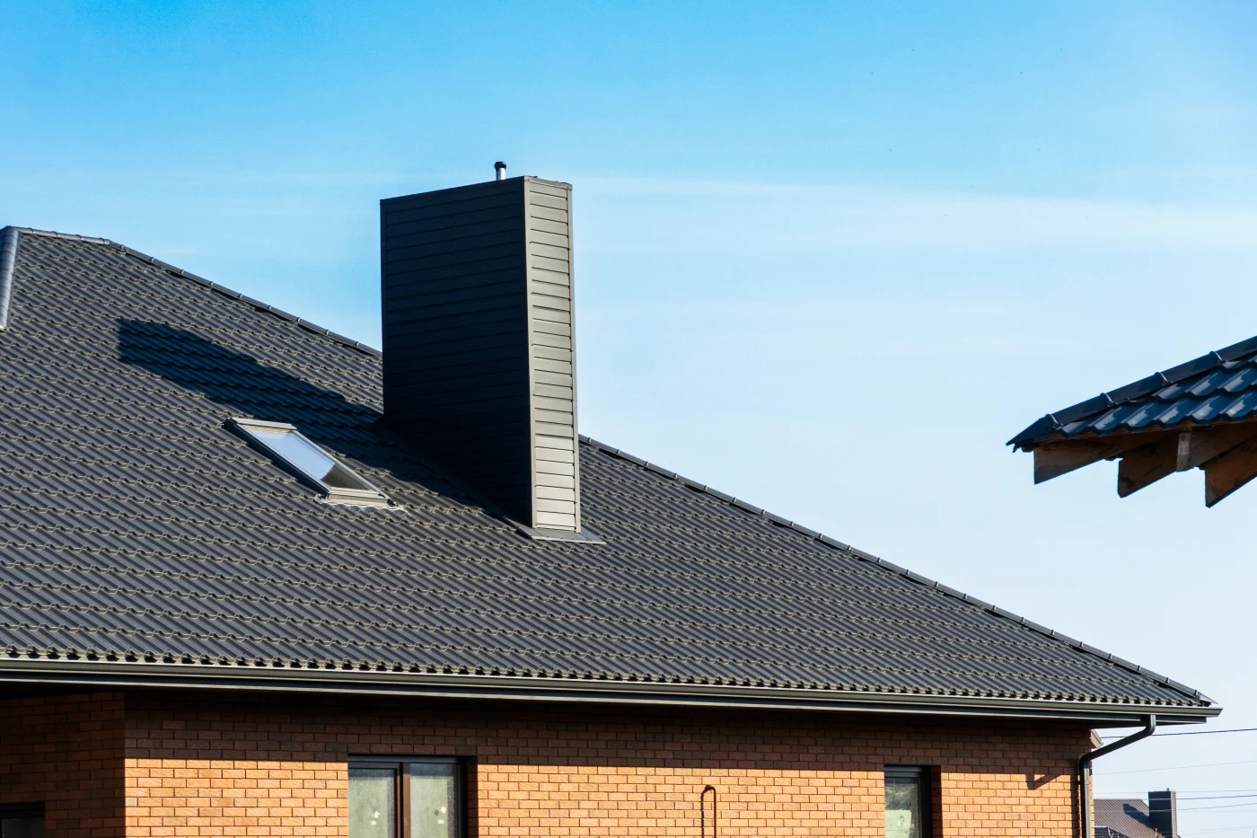 A metal tile roof with a brown chimney on a brick house.