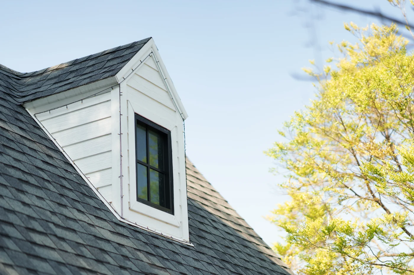 A close-up view of a white dormer, black window, and asphalt shingle roof.