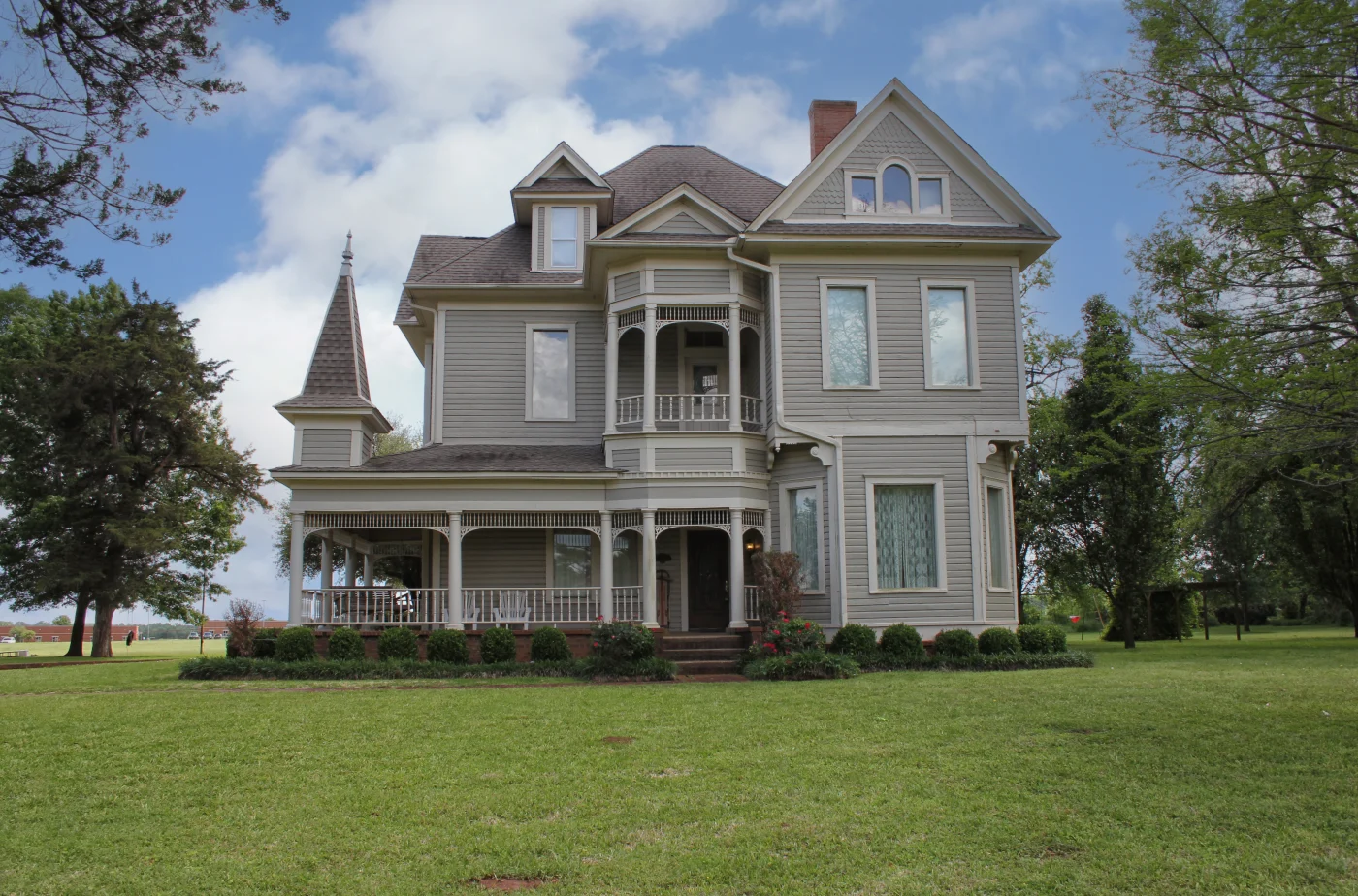 A multi-story gray house with a white porch, a small balcony, white windows, and an asphalt shingle roof.