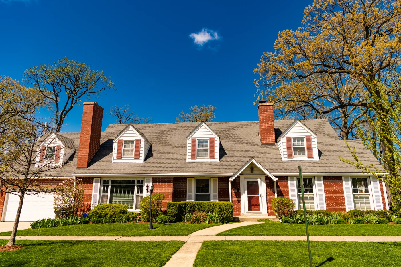 A brick house with white windows, white shutters on the first floor, brown shutters on the second floor, a white garage door, two brick chimneys, and a large asphalt shingle roof.