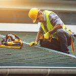 A roofing contractor wearing a hard hat and safety vest uses a power drill to secure materials during a professional roof replacement project.