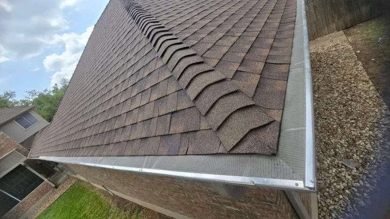 A close-up view of a residential brick home shows newly installed brown asphalt shingles fitted with a seamless gutter installation along the roofline.