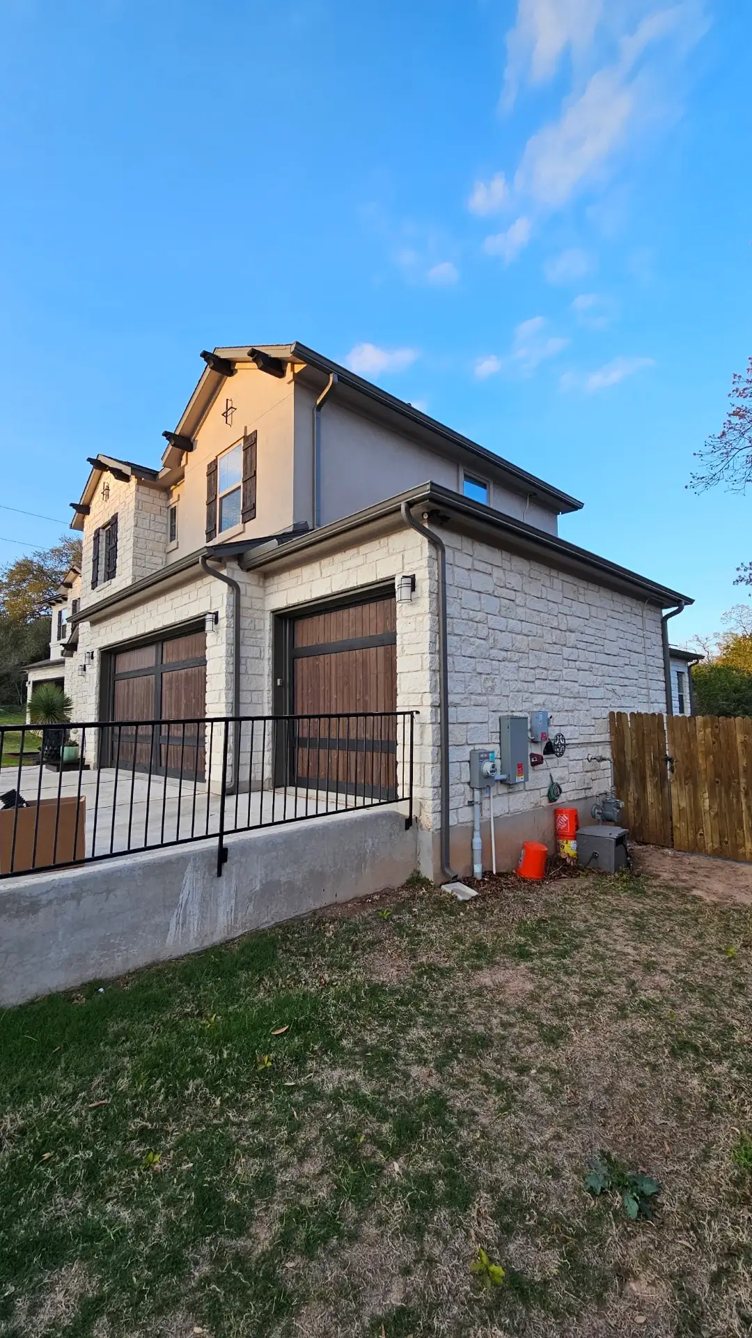 The corner of a stone and stucco house with a black fence, wooden garage doors, white windows with wooden shutters, brown gutters, and a brown downspout.