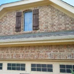 A close-up look at a brick house with an asphalt shingle roof, a tan garage with windows at the top, and a window above the garage with wooden shutters.