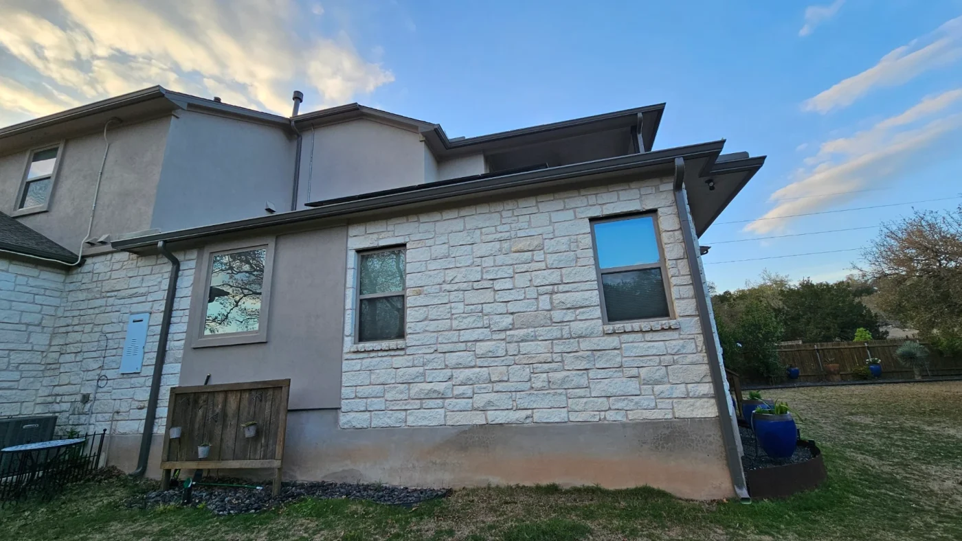 The side of a stone and stucco house with multiple windows, brown gutters, and a brown downspout.