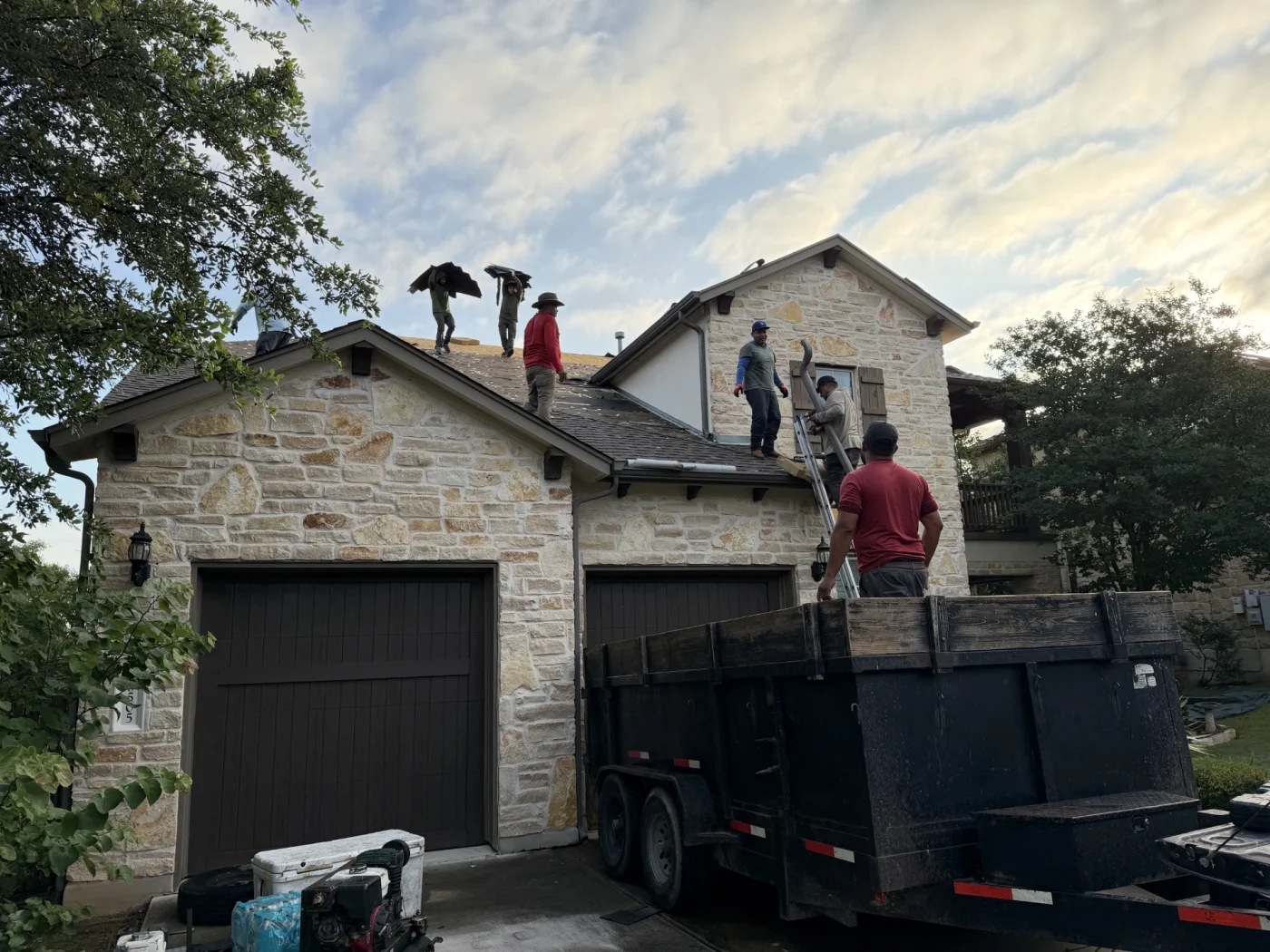 Roofers replacing the asphalt shingle roof of a stone house with wooden garage doors.