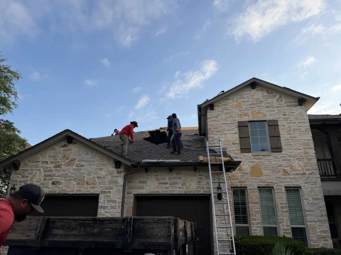 Roofers replacing the asphalt shingle roof of a stone house with windows and wooden shutters.
