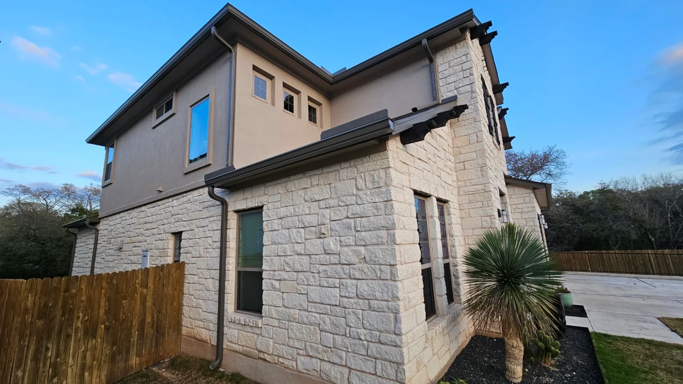 The corner of a stone and tan stucco house with brown windows, brown gutters, a brown downspout, and a wooden fence.