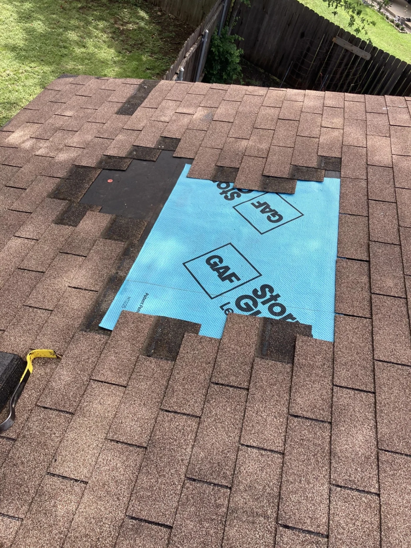 A section of brown asphalt shingles has been removed from a residential roof to expose the blue underlayment as part of a targeted roof repair project.