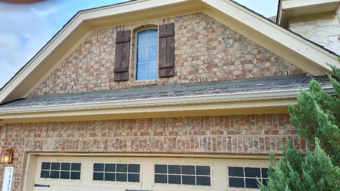 An upper gabled section of a home features a detailed brick facade surrounding a window with dark wood shutters, showcasing a structure that's benefited from professional masonry repair services.