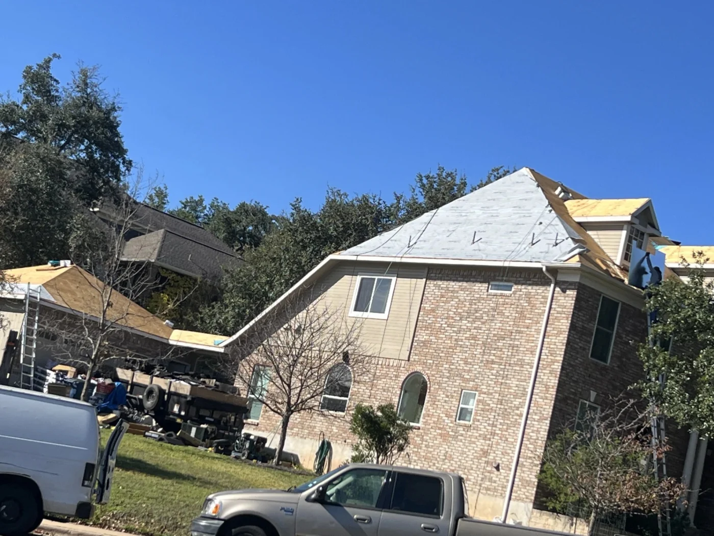 Work trucks sit parked outside a large brick home where the multi-gabled roof has been stripped down to the decking and underlayment for professional roofing services.